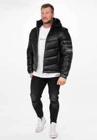 Zwarte pufferjack met een capuchon, rits aan de voorkant en een gestructureerde afwerking. Voorzien van zijzakken en een patch op de mouw. Draag het over een wit t-shirt en zwarte jeans.