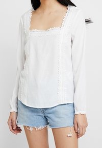 Blouse blanche à manches longues avec un décolleté carré, ornée de dentelle et de détails texturés sur le devant. Assortie à un short en denim effiloché.