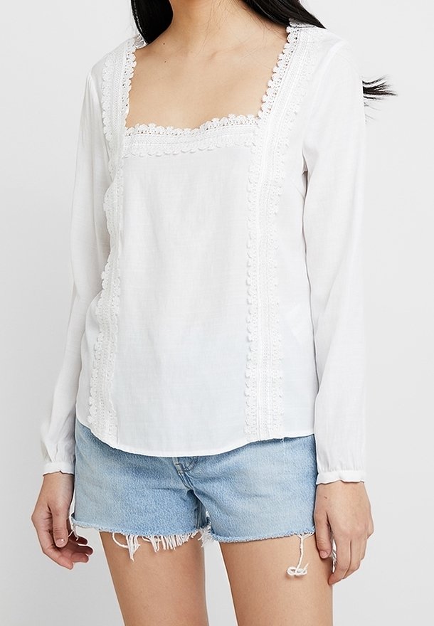 Blouse blanche à manches longues avec un décolleté carré, ornée de dentelle et de détails texturés sur le devant. Assortie à un short en denim effiloché.