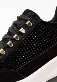 Svart atletisk sneaker med en texturerad mockaövre, glänsande nitar, metalliska öglor och en vit sula. Snörning i grått.