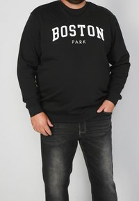 Černá mikina s nápisem "BOSTON PARK" bílým písmem přes hrudník. Dlouhé rukávy, kulatý výstřih, kombinovaná s tmavými džíny a texturovanou látkou.