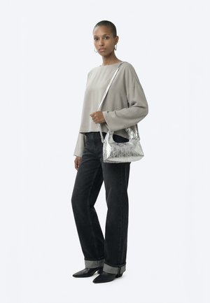 Ženska, ki nosi ohlapno bež majico, črne zavihane kavbojke, črne koničaste škornje in nosi svetlečo srebrno torbico čez ramo na enobarvnem ozadju.