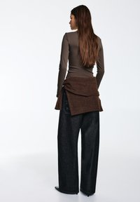 Top marrone a maniche lunghe abbinato a pantaloni di jeans neri a gamba larga. Un maglione marrone è annodato in vita. Texture lisce e design casual.