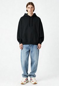 Čierna mikina s kapucňou a predným vreckom, skombinovaná so svetlomodrými voľnými džínsami a bielymi teniskami s červenými a modrými akcentmi.