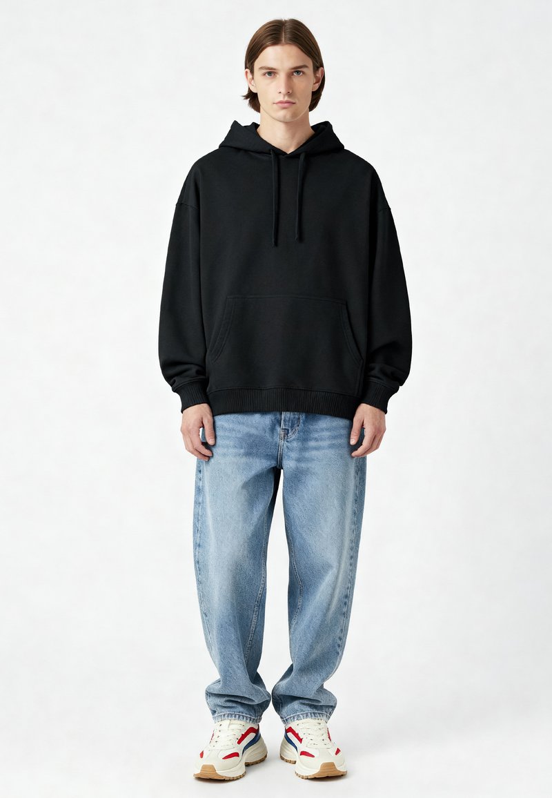 Čierna mikina s kapucňou a predným vreckom, skombinovaná so svetlomodrými voľnými džínsami a bielymi teniskami s červenými a modrými akcentmi.