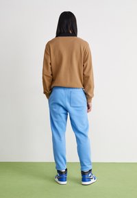 Bruine langesleeve top, van textuurstof, gecombineerd met lichtblauwe trainingsbroek. Zwarte accenten op de manchetten, met blauwe en witte sportschoenen.
