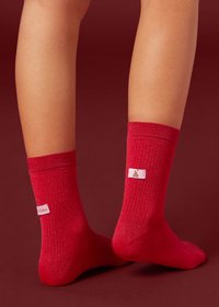 Chaussettes rouges côtelées avec un léger éclat, ornées d'une étiquette rose avec un petit motif. Design à longueur de cheville, texture douce, sans motifs apparents.