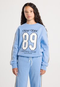 Girl with long curly hair wears a blue sweatshirt and matching pants with white and black stripes, standing against a plain background.