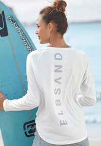 White long-sleeve shirt with a crew neck, featuring the text "ELBSAND" in light grey on the back. Smooth, lightweight fabric. Surfboard nearby.