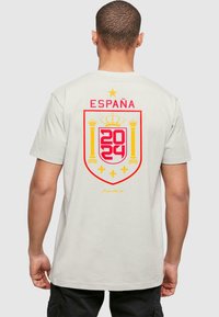 Beige bomull T-skjorte med et rødt og gult spansk emblem med "ESPAÑA" og "2024" på baksiden, detaljert med kroner og liljer.