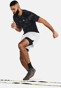 Zwarte camouflage sportshirt, witte shorts met zwarte accenten, grijze trainingsschoenen, man die halverwege een sprongetje op een agility ladder maakt, gespierd postuur.