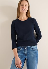 Haut bleu marine ajusté à manches longues en tissu côtelé, associé à un jean en denim bleu clair, avec une taille classique et des poches frontales.