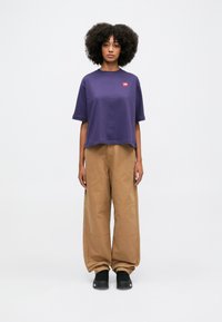 Maglietta cropped viola con logo rosso, abbinata a pantaloni beige larghi e scarpe nere. Texture liscia con dettagli di design semplici.