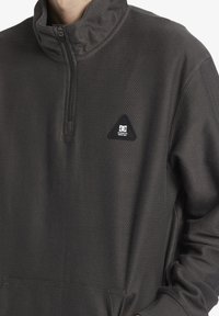 Dark gray pullover with a zippered neck, textured fabric, a triangular logo patch on the chest, and kangaroo pocket at the front.