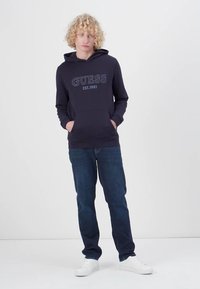Felpa blu marina con il logo "GUESS EST. 1981", tasca frontale e cappuccio regolabile; abbinata a jeans scuri e scarpe da ginnastica bianche.