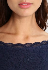 Haut en dentelle bleu marine avec un col en forme de festons, présentant des motifs floraux et une finition texturée, mettant en valeur des détails en dentelle intricats.