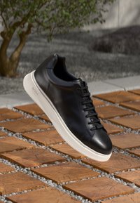 Černá kožená teniska s hladkou texturou, zaobleným tvarem, šněrováním a bílou gumovou podrážkou. Logo značky je jemně zobrazeno na straně.