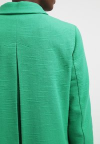 Zelený blazer vyrobený z texturované tkaniny, s vystouplým límcem a ventilačním otvorem na zadní straně, se viditelným stehem podél švů.