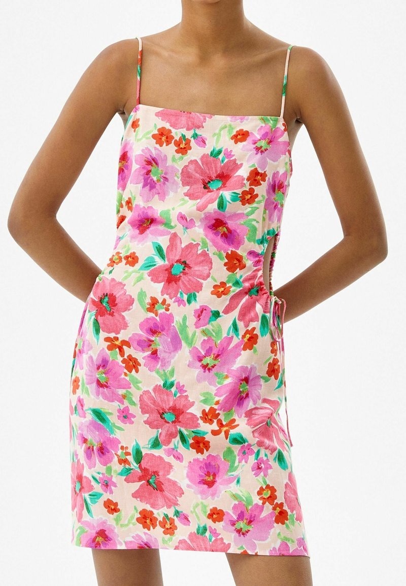 Vestido floral con tirantes finos, que presenta un patrón rosa y verde sobre un fondo claro. La fruncido lateral añade textura y detalle.