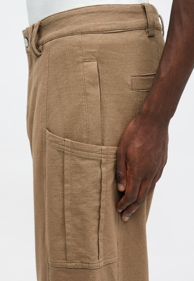 Bēšas krāsas kokvilnas kargo bikses ar vaļēju piegriezumu, tiem ir sānu kabatas un pogu aizdare pie vidukļa, parādītas sānskatā.