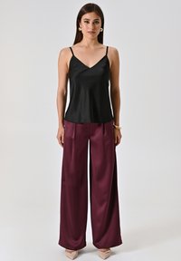 Camisola nera in satin con spalline sottili abbinata a pantaloni ampi in satin color bordeaux. L'insieme presenta una texture liscia e una vestibilità rilassata.