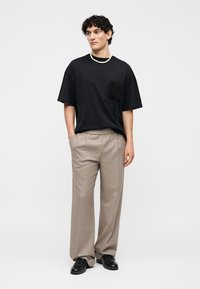 Jeune homme aux cheveux bouclés portant un t-shirt noir oversize avec une poche, un pantalon beige à jambes larges, des chaussures noires et un collier de perles blanc.