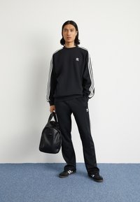 Svart idrottströja med vita ränder, matchande svarta byxor och svarta sneakers. Modellen håller en svart väska. Logotyp på bröstet.