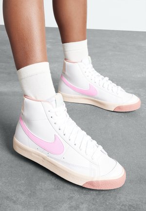 Jambes portant des baskets montantes blanches avec des logos swoosh roses, des semelles beige et des chaussettes blanches, debout sur un sol gris.