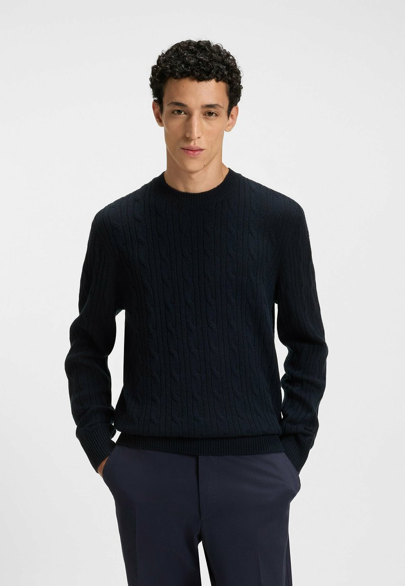 Navy-blauer Strickpullover mit geripptem Rundhalsausschnitt und langen Ärmeln, aus weichem, strukturiertem Stoff. Kombiniert mit dunklen Hosen.