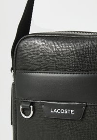 Sort tekstureret læder Lacoste skuldertaske med sølvmetallisk ring og synlig lynlåsdetalje.