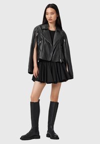Svart läderbikerjacka med silverblixtlås, lager-på-lager över en svart utsvängd kjol. Modellen bär knähöga svarta stövlar. Slät textur.