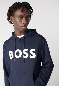 Sweatshirt azul-marinho com um grande logótipo branco "BOSS". Apresenta um capuz com cordão e bolso frontal. Tecido de algodão macio com punhos canelados.