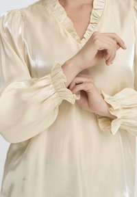 Lys beige bluse med V-hals, der har blødt rynkede detaljer på kraven og ærmerne. Fremstillet af et silkeagtigt, glat materiale med en subtil glans.