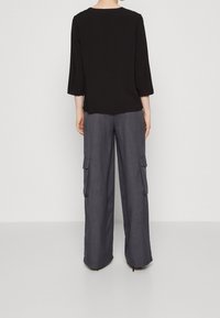 Person in einem schwarzen Langarmoberteil und lockeren grauen Cargo-Hosen mit Seitentaschen, steht vor einem schlichten weißen Hintergrund, mit dem Rücken zur Kamera.