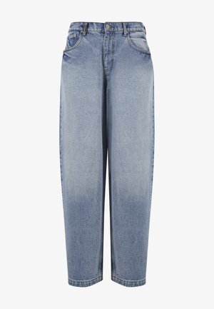 Person trägt hellblaue Jeans mit weitem Bein, weiße Sneakers und ein weißes Tanktop, stehend vor einem einfarbigen grauen Hintergrund.