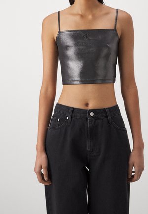 Femme portant un débardeur court noir brillant à fines bretelles et un jean noir ample taille haute sur un fond uni.