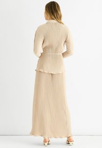 Beige pleated dress with a peplum waist, long sleeves, and a flowing skirt. Features subtle texture and a slit at the back.