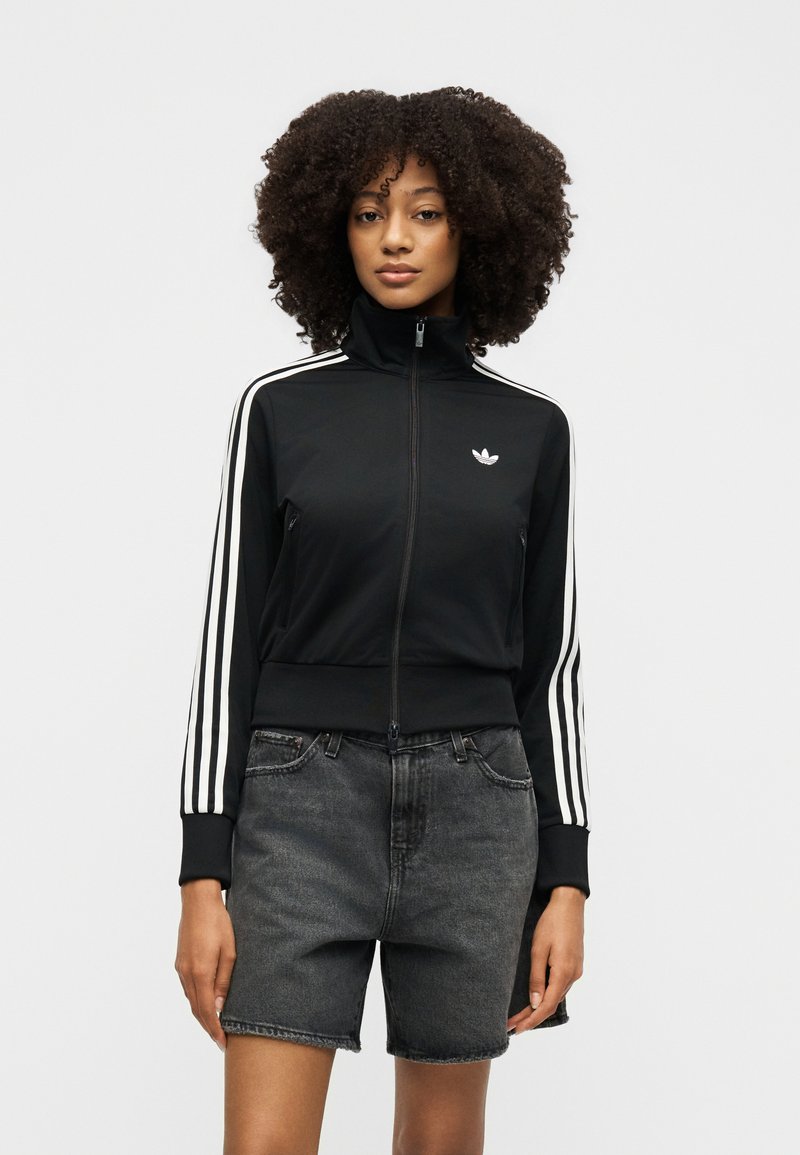 Femme aux cheveux bouclés portant une veste zippée noire Adidas avec des bandes blanches et un short en jean gris foncé, debout devant un fond uni.