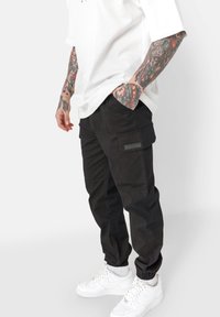 Zwarte cargobroek met elastische boorden, voorzien van meerdere zakken en een gestructureerd oppervlak. Draagt een witte oversized t-shirt en witte sneakers.