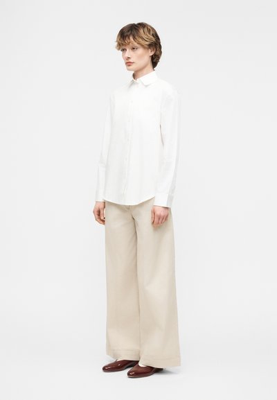 Άτομο που στέκεται φορώντας λευκό πουκάμισο με κουμπιά, μπεζ παντελόνι με φαρδιά μπατζάκια και καφέ παπούτσια μπροστά σε απλό ανοιχτό φόντο.