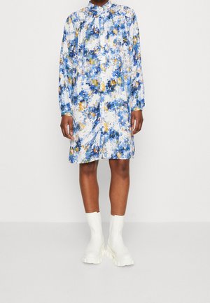 Frau trägt ein locker sitzendes, langärmeliges, knielanges Kleid mit blau-gelben Blumenmustern, kombiniert mit weißen, klobigen Stiefeletten.