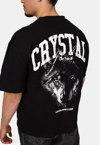 Černé bavlněné tričko s velkým grafickým motivem vlka a textem "CRYSTAL Paris" v tučném bílém písmu na zádech. Casual střih.