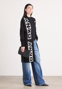 Črn kardigan z belim logotipom DKNY, dolgi rokavi in odprt sprednji del. Usklajen z modrimi širokimi hlačami in črno torbico na roko z logotipom.