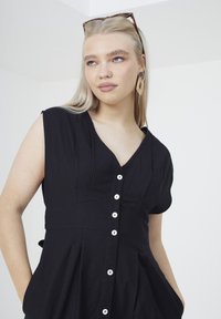 Vestito nero a maniche corte con scollatura a V, chiusura con bottoni e gonna plissettata. Tessuto liscio con tasche su entrambi i lati.