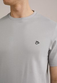 Hallhalline lühikeste põlvede käistega t-särk, mis on valmistatud siledast puuvillast. Vasakul rinnal on väike must tikandiga linnu logo. Lihtne ümar kaelus.