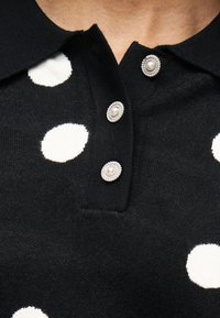 Μαύρο πλεκτό τοπ με μεγάλα λευκά πουά, διακοσμητικά κουμπιά με κέντρο από πέρλες και γιακά με V-σχήμα λαιμόκοψη.