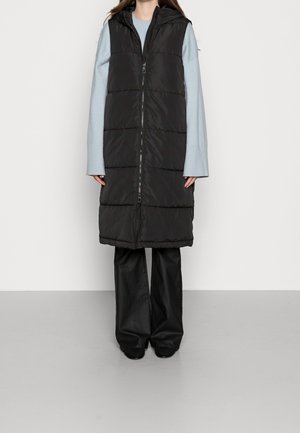 Personne portant une longue doudoune noire avec capuche, un pull à manches longues bleu clair et un pantalon large noir, debout contre un mur uni.