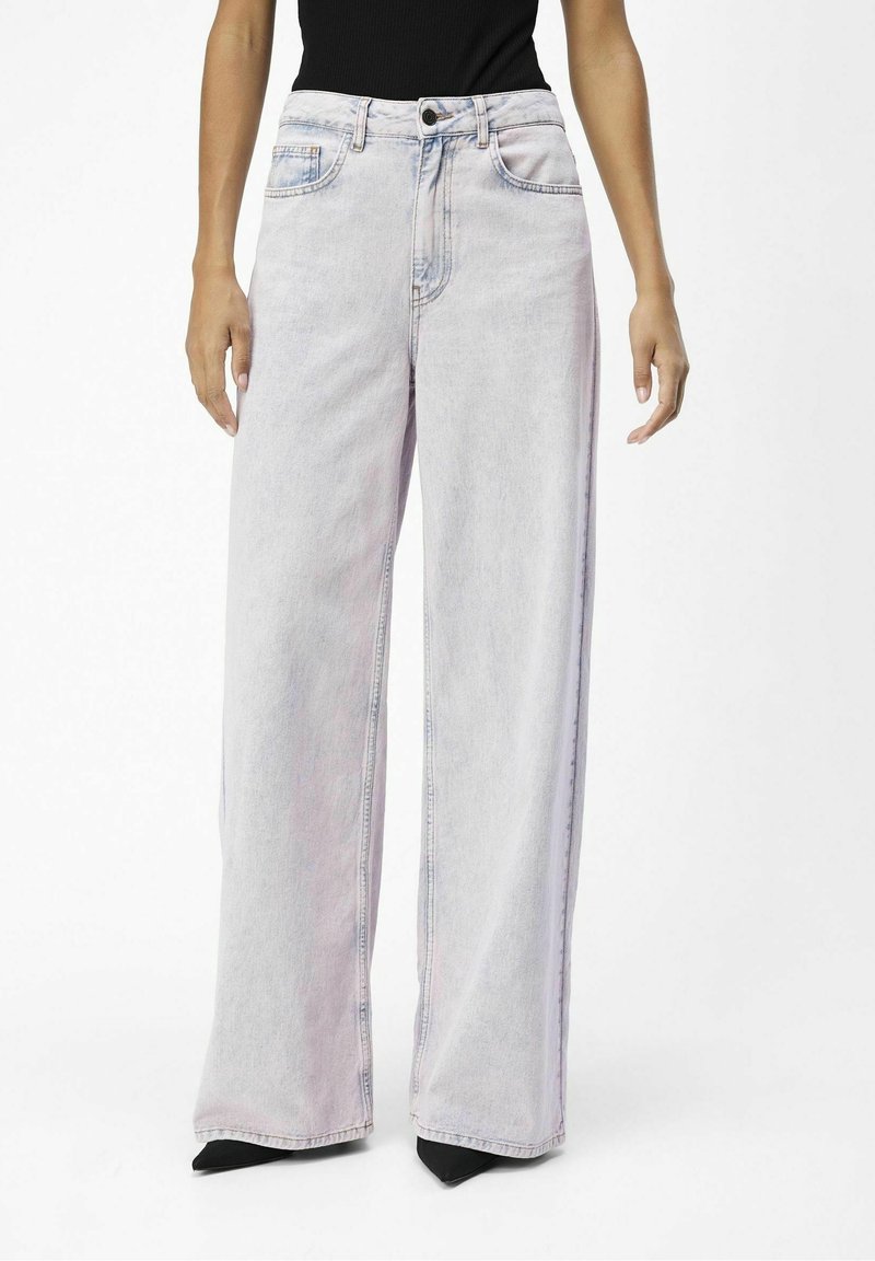 Persona que lleva puestos jeans de tiro alto, en un lavado claro y de pierna ancha, combinados con zapatos negros de punta, sobre un fondo liso.