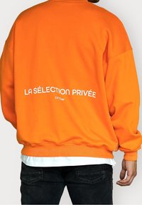 Felpa arancione con polsini e orlo a coste. Presenta la scritta bianca "LA SELECTION PRIVÉE ESTIM" stampata sul retro. Tessuto morbido.