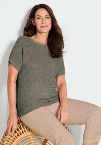 Grijze gebreide top met korte mouwen, met een textuurpatroon; gecombineerd met lichtbeige broek. Model zit op een gevlochten rotan stoel.